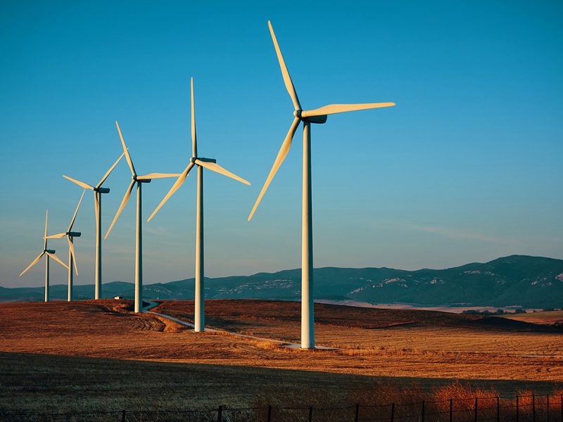 Six electric wind turbines in a row located in grassy field with mountains in background